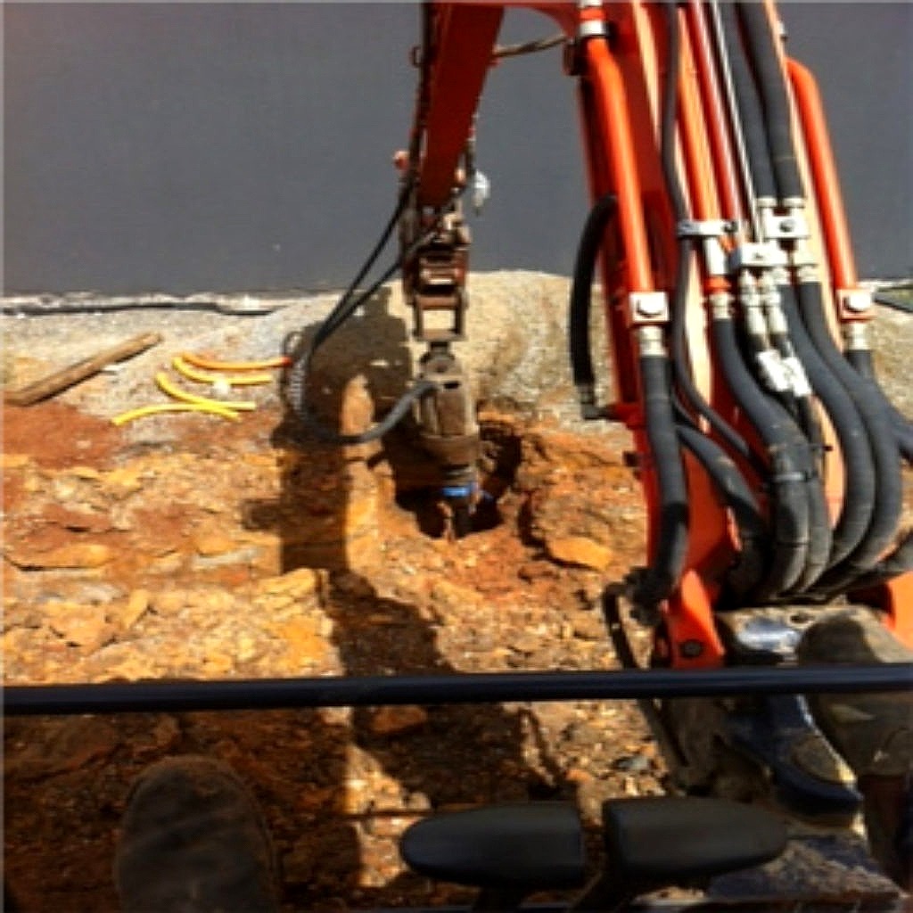 Hydraulic rock breaker attachment on a mini excavator splitting rock along an open trench line