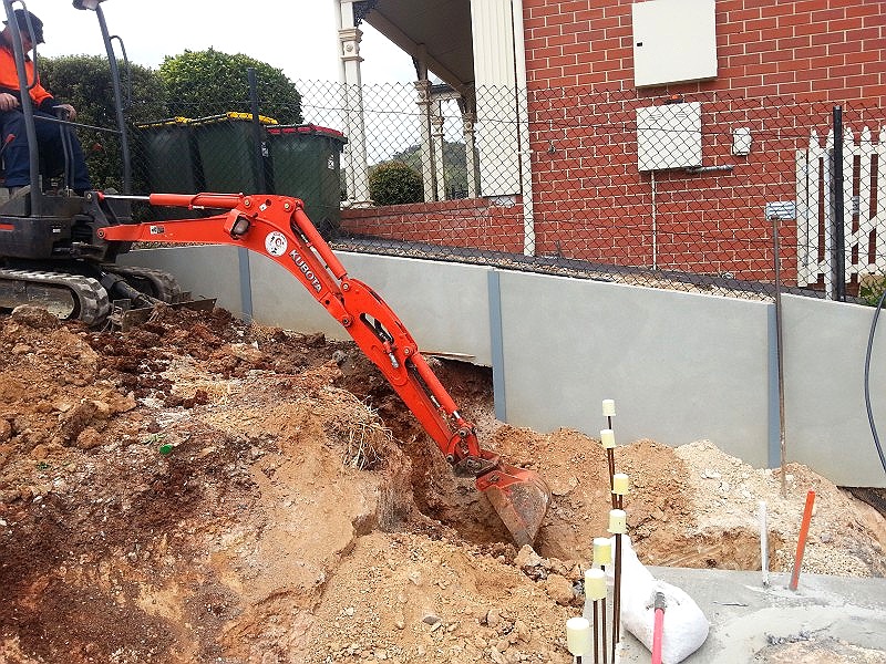 Orange Kubota mini excavator digging beside a rendered retaining wall at a brick home, with electrical conduit stakes marked out