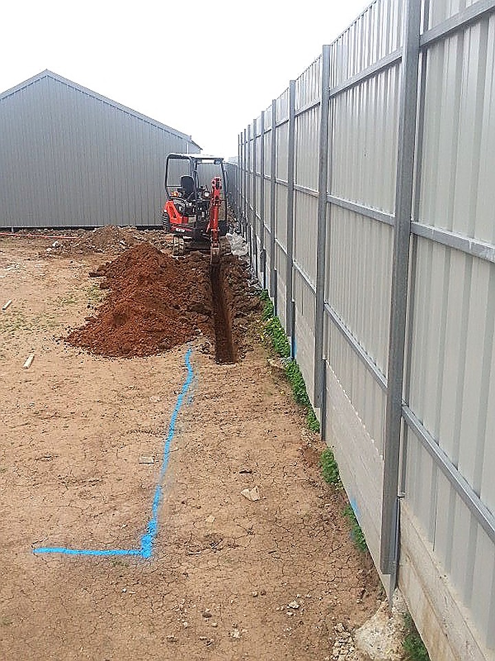 Mini excavator cutting a service trench along a Colorbond fence line, with blue Dial Before You Dig service marking sprayed on the ground